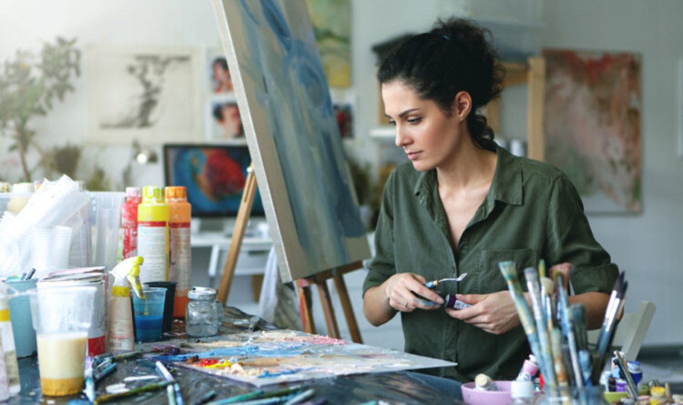 young-female-painter-sitting-table-surrounded-with-various-paint-brushes-watercolors-while-creating-beautiful-picture-easel-creative-wo